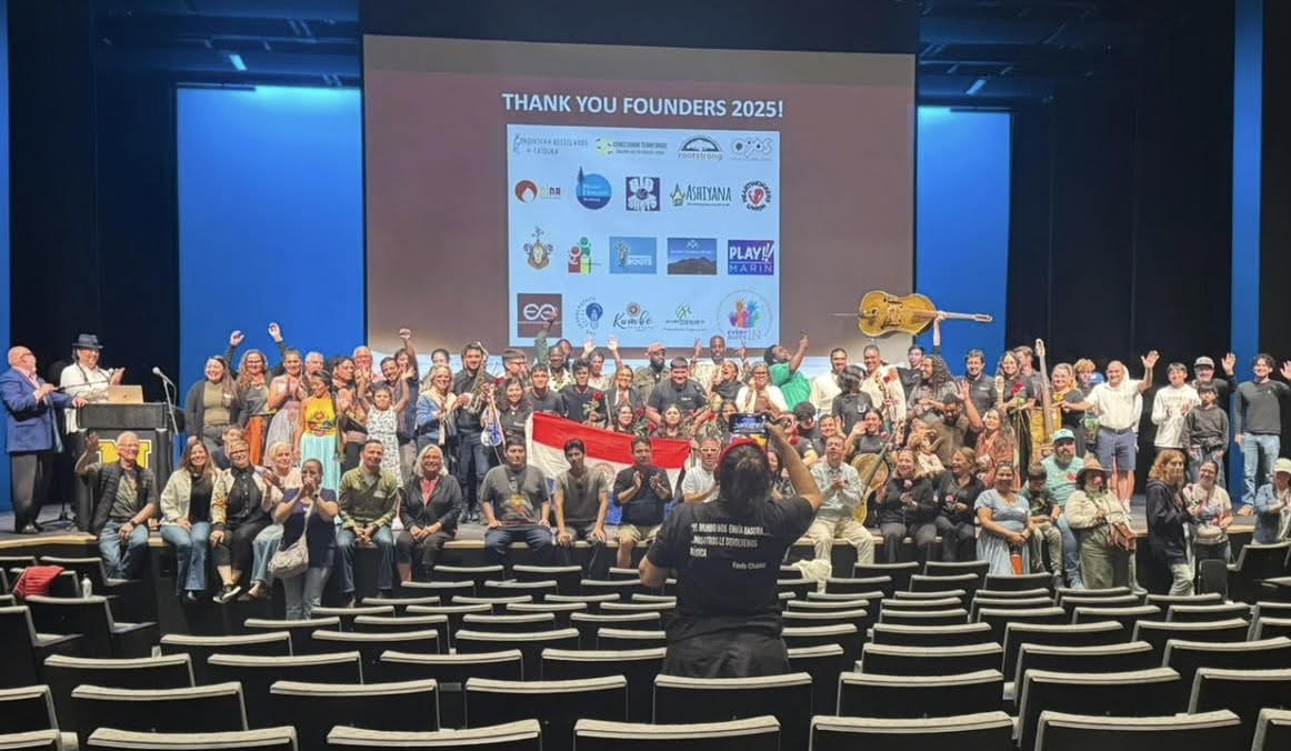 A large group gathers onstage beneath a screen thanking the 2025 founders, with musical instruments and raised hands.